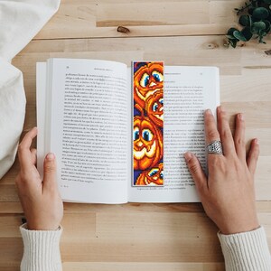 May include: A close-up of a book with a bookmark featuring a colorful pixelated design of several orange pumpkins with smiling faces. The bookmark is placed between the pages of the book.