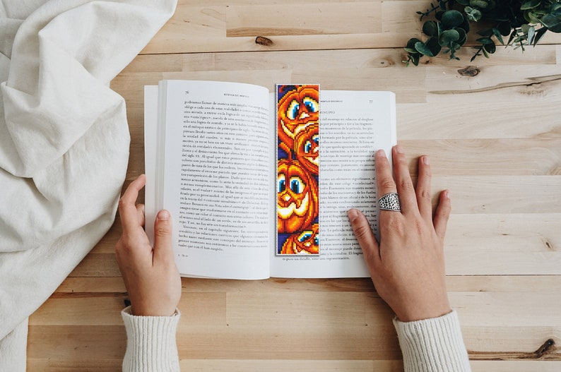 May include: A close-up of a book with a bookmark featuring a colorful pixelated design of several orange pumpkins with smiling faces. The bookmark is placed between the pages of the book.