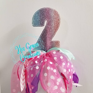 May include: A glittery rainbow number two cake topper with pink, purple, and white polka dot ribbons.