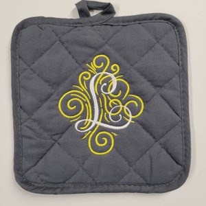 Puede incluir: Un posavasos acolchado gris con un diseño bordado amarillo que presenta una letra L estilizada.
