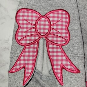 May include: Close-up of a gray sweatshirt sleeve featuring a large, pink gingham bow applique. The bow has a bright pink outline and a white and pink checkered pattern. The bow is centered on the sleeve.
