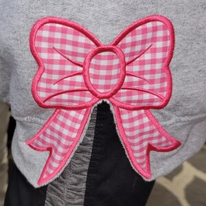 May include: A gray sweatshirt features a large, pink gingham bow applique. The bow has a pink outline and a white and pink checkered pattern. The bow is centered on the sweatshirt, adding a decorative touch to the garment.