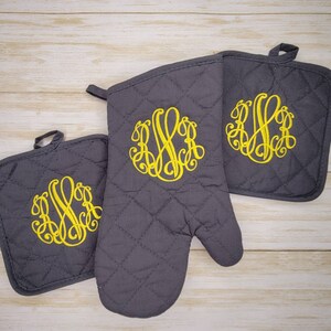 May include: A set of three grey quilted oven mitts with yellow embroidered monograms. The mitts are designed to protect hands from heat while cooking or baking.