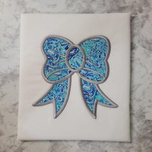 May include: A white fabric square featuring a large embroidered bow. The bow has a marbled pattern in shades of blue and teal, outlined with a silver-gray border. The design is centered on the fabric.