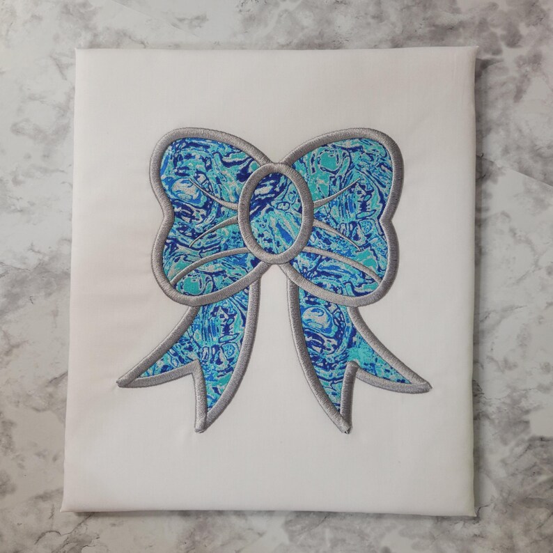 May include: A white fabric square featuring a large embroidered bow. The bow has a marbled pattern in shades of blue and teal, outlined with a silver-gray border. The design is centered on the fabric.