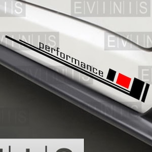Op de afbeelding: Witte auto met zwarte en rode "performance" stickers. De stickers tonen het woord "performance" in zwarte letters, met een ontwerp van zwarte, rode en witte strepen. De afbeelding bevat ook de tekst "EVINIS".