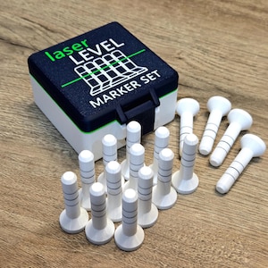 Op de afbeelding: Een laser waterpas markerset met een marineblauwe en witte doos en witte markers. De doos heeft de woorden "laser LEVEL MARKER SET" in groen en wit. De markers zijn wit met zwarte strepen en een afgeronde bovenkant.