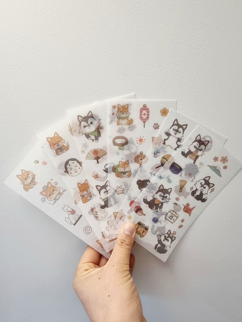 May include: A set of six sheets of clear stickers featuring illustrations of cartoon dogs in various poses and wearing traditional Japanese clothing. The stickers also feature other Japanese themed illustrations such as a fan, a lantern, a mountain, and a teacup. The stickers are on a white background.