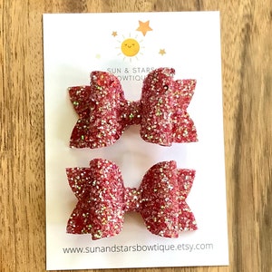 May include: Two red glitter hair bows with a white background. The bows are made of a sparkly material and are attached to a clip. The text "SUN & STARS BOWTIQUE" is printed on the background.