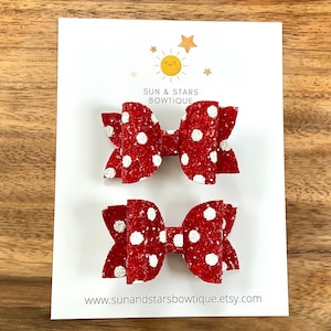 May include: Two red and white polka dot hair bows with glitter. The bows are on a white card with the text "Sun & Stars Bowtique" and a website address.