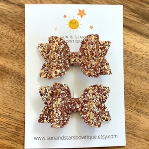 Puede incluir: Dos lazos para el pelo de purpurina dorada en una tarjeta blanca con el texto "Sun & Stars Bowtique" y una dirección de sitio web.