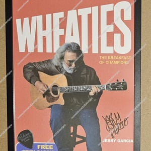 May include: Framed print featuring a vintage Wheaties box design with Jerry Garcia playing a guitar. The box art includes the text "Whole Grain Flakes" and "The Breakfast of Champions." Includes a free Jerry Garcia guitar pick.