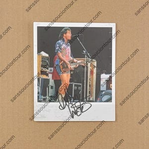 May include: Polaroid photo of a musician playing a mandolin on stage. The musician wears a patterned shirt and denim shorts. The photo is signed in black ink. The background includes speakers and stage equipment.