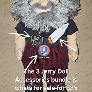 May include: A plush doll resembling a man with a gray beard, glasses, and a black t-shirt. The doll wears burgundy corduroy pants and black sneakers. Accessories include a peace sign pendant, a badge, and a paper cutout of a lit cigarette.