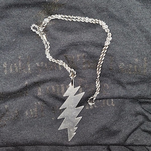 May include: Silver-colored necklace with a lightning bolt pendant. The pendant is a stylized zig-zag shape. The chain is a twisted rope design. The necklace is displayed on a dark, textured surface.