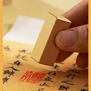 Pode incluir: Um carimbo retangular dourado com bordas arredondadas. O carimbo está sendo segurado por uma mão e está prestes a ser pressionado em um pedaço de papel com caracteres chineses escritos nele. O carimbo tem uma impressão vermelha de um caractere chinês no papel.