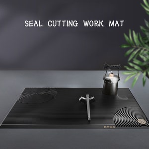 May include: Black seal cutting work mat with a circular pattern and lines. A small black metal incense burner and a metal tool are on the mat. The text "SEAL CUTTING WORK MAT" is at the top of the image. The text "东洋出品" is at the bottom of the image.