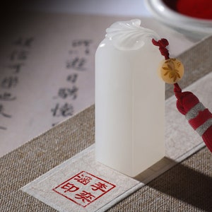 Pode incluir: Selo de jade branco com um design de flor esculpido no topo. O selo tem uma borla vermelha com uma conta dourada e uma seção listrada vermelha e branca. O selo está apoiado em um pedaço de papel com um carimbo vermelho que diz "印章".