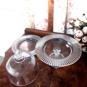 May include: A clear glass butter dish with a domed lid and a matching base plate. Both pieces feature a distinct vertical ribbed pattern along the edges. The lid has a small, round knob handle at the top, and the base plate has a central circular indentation.