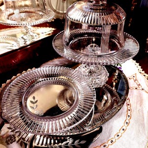 May include: A clear glass cake stand with a matching domed lid featuring vertical ribbed detailing. A separate clear glass plate with a smooth centre and a ribbed rim sits below, decorated with a delicate etched leaf and flower pattern.