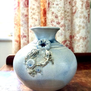 May include: A pale blue ceramic vase with a rounded form and a flared neck. It has a raised floral design and a dark blue rim. This decorative vase is a home decor item, suitable for displaying flowers.