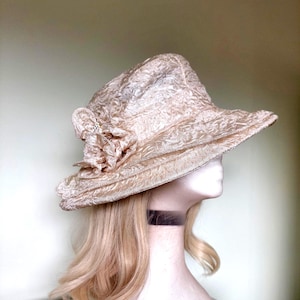 May include: A champagne-coloured hat with a textured, crinkled fabric finish. The hat features a wide, slightly downturned brim and a decorative fabric flower accent on the side, which is adorned with a small, clear, sparkling crystal detail.