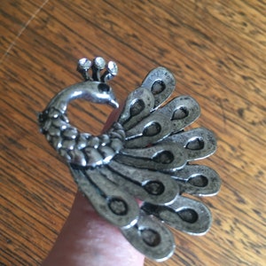 May include: A silver peacock ring with clear rhinestones on the head. The peacock's tail feathers are spread out in a fan shape.