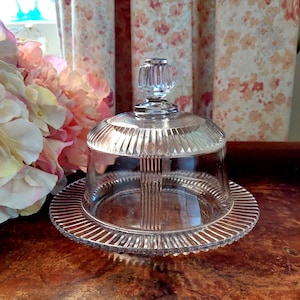 May include: A clear glass cloche with a ribbed design, featuring a domed lid and a circular base. The lid has a decorative, faceted knob. The cloche sits on a wooden surface, with floral curtains and pink flowers in the background.