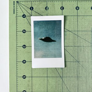 Puede incluir: Una foto borrosa de un OVNI gris oscuro contra un cielo azul. La foto est&aacute; impresa sobre un fondo blanco y est&aacute; enmarcada por un borde blanco.