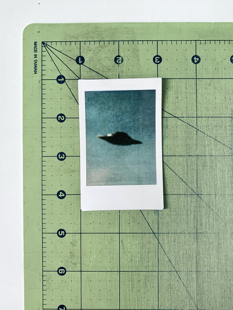 Puede incluir: Una foto borrosa de un OVNI gris oscuro contra un cielo azul. La foto est&aacute; impresa sobre un fondo blanco y est&aacute; enmarcada por un borde blanco.