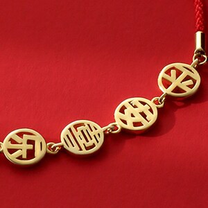Peut inclure: Un bracelet en or avec un cordon rouge et quatre breloques circulaires représentant des caractères chinois. Les breloques sont reliées par de petits maillons en or.