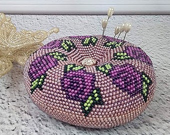 May include: A pink and purple beaded pincushion with a flower design. The pincushion has a circular shape and is filled with pins.