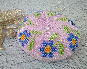 May include: A pink beaded pincushion with a pearl button in the center and blue and yellow flowers around the edge.  The pincushion has several white pearl-headed pins sticking out of the top.