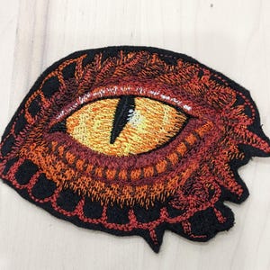 Peut inclure: Patch brodé représentant un œil de dragon rouge avec une pupille noire et un iris orange. L'œil est entouré d'une bordure noire et rouge.
