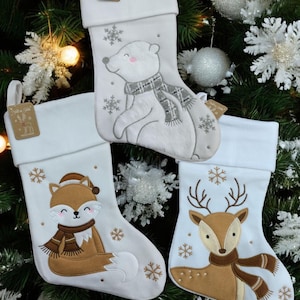 Op de afbeelding: Drie witte kerstsokken met dierenontwerpen. Eén sok heeft een ijsbeer met een sjaal, een andere een vos met een hoed en sjaal, en de derde een rendier met een sjaal. Elke sok heeft sneeuwvlokaccenten.