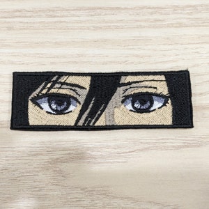 Peut inclure: Patch brodé représentant un gros plan sur une paire d'yeux avec des cheveux noirs. Les yeux sont bruns avec des cils noirs et des pupilles blanches. Les cheveux sont noirs et couvrent une partie des yeux.