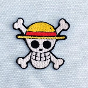 Peut inclure: Écusson brodé représentant une tête de mort blanche avec des yeux noirs et une bouche souriante, coiffée d'un chapeau de paille jaune avec un bandeau rouge. Deux tibias croisés blancs sont derrière le crâne.