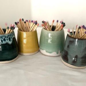 May include: Four ceramic match holders in teal, yellow, and green, each filled with wooden matches. The holders have a glossy finish and a white base. The match heads are various colours, including pink, purple, and orange.