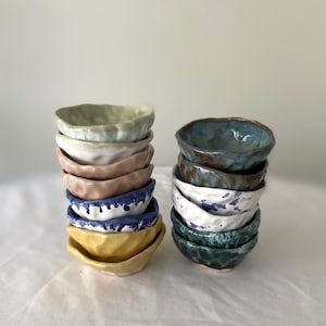 May include: Two stacks of small, handmade ceramic bowls in various colours. The bowls have irregular, textured edges and a variety of glazes, including blue, yellow, white, and green. The bowls are stacked on a white surface.