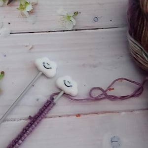 May include: Knitting needles with white cloud-shaped tops and smiling faces are inserted into a row of purple stitches. A skein of variegated yarn in shades of purple, brown, and cream is visible in the background. The image is set on a white wooden surface.