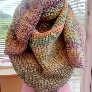 May include: A hand-knitted cowl in a mix of pink, yellow, green, and grey. The scarf is displayed on a pink mannequin. The cowl has a ribbed texture and a folded design, highlighting the colour variations.