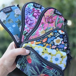 May include: A collection of five zipped pouches in various floral and botanical prints, each featuring a black trim and a black fabric tag labelled KK Studio Australia. The pouches display patterns including blue, pink, and black backgrounds.