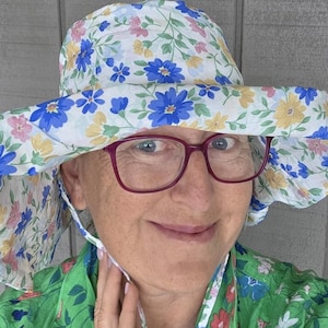 May include: A wide-brimmed sun hat featuring a white base with a floral pattern of blue, yellow, and pink flowers. The hat includes a chin strap and is worn with dark red-rimmed spectacles. The fabric has a soft, lightweight appearance.