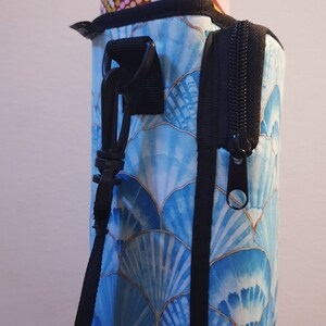 K&ouml;nnte beinhalten: Ein blauer und goldener Wasserflaschenhalter mit Rei&szlig;verschlussfach und schwarzem Gurt. Der Halter hat ein Muster aus blauen und goldenen Muscheln.