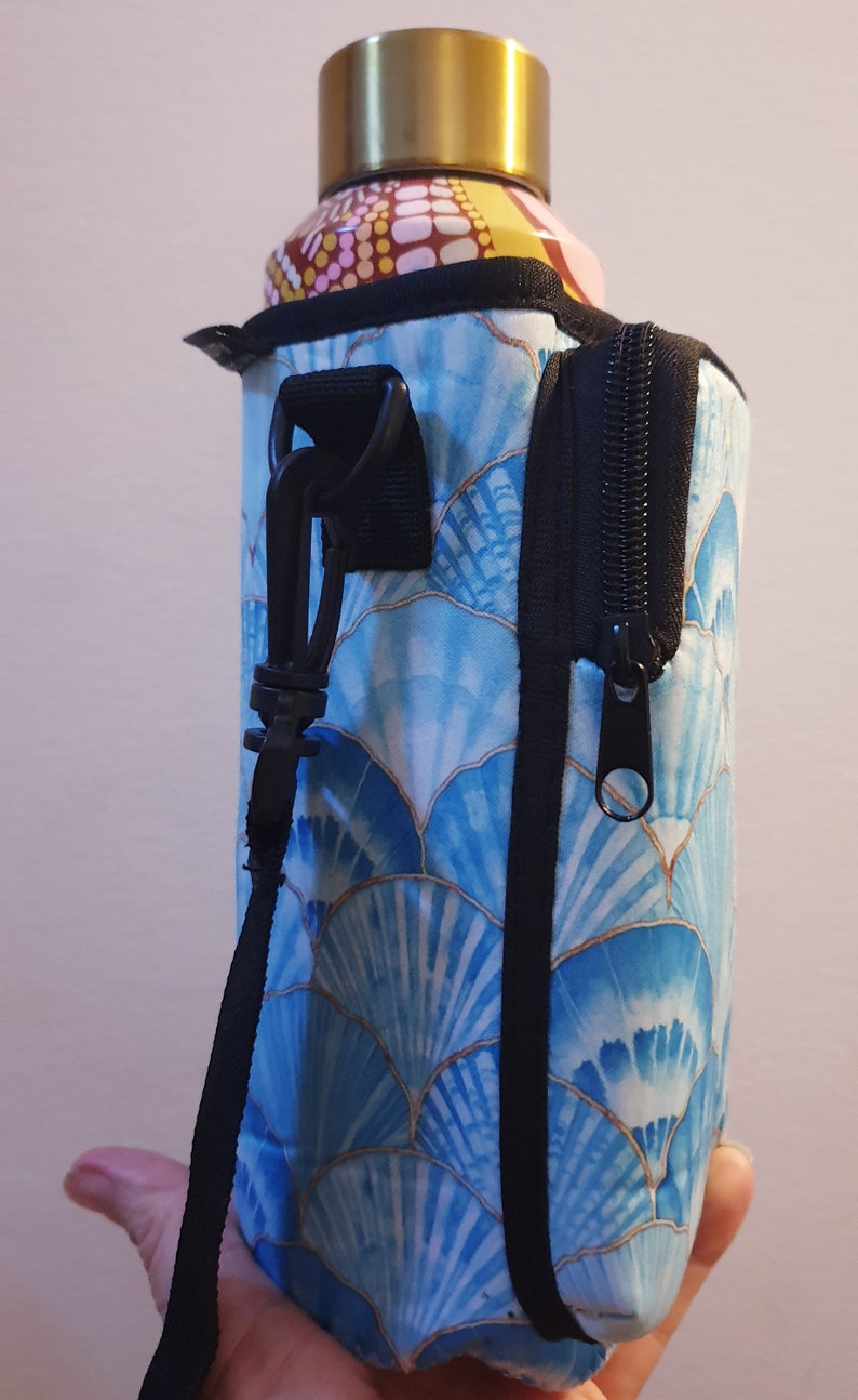 K&ouml;nnte beinhalten: Ein blauer und goldener Wasserflaschenhalter mit Rei&szlig;verschlussfach und schwarzem Gurt. Der Halter hat ein Muster aus blauen und goldenen Muscheln.