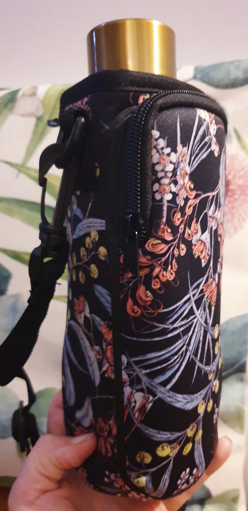 K&ouml;nnte beinhalten: Schwarze isolierte Wasserflaschenh&uuml;lle mit einem Blumendruck mit rosa, blauen und gelben Blumen. Die H&uuml;lle hat einen Rei&szlig;verschluss und einen schwarzen Gurt.