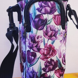 K&ouml;nnte beinhalten: Ein isolierter Flaschenhalter mit floralem Druck in Schwarzwei&szlig;, Rei&szlig;verschluss und verstellbarem Trageriemen. Der Halter hat einen wei&szlig;en Hintergrund mit violetten und rosafarbenen Tulpen.