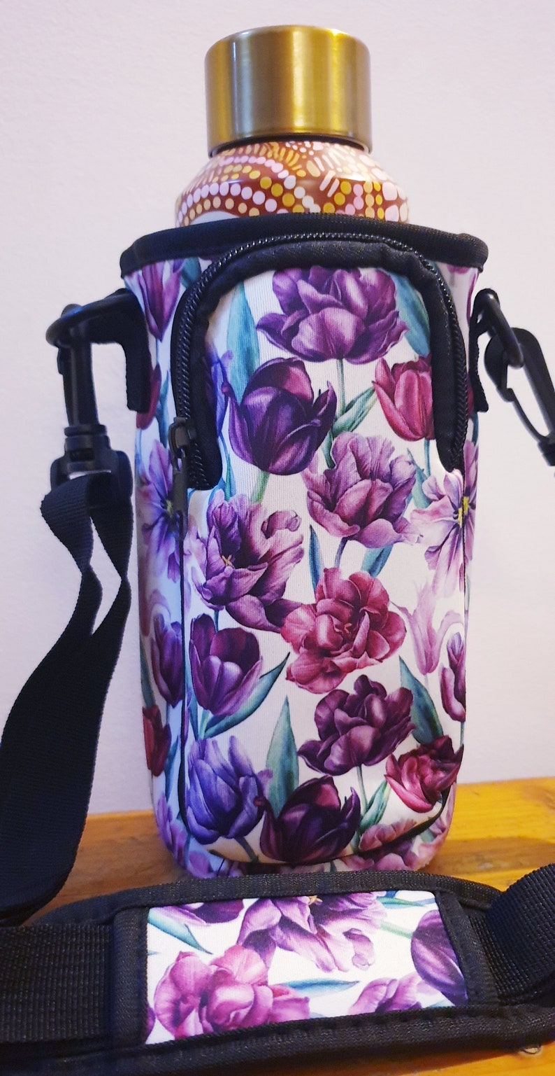 K&ouml;nnte beinhalten: Ein isolierter Flaschenhalter mit floralem Druck in Schwarzwei&szlig;, Rei&szlig;verschluss und verstellbarem Trageriemen. Der Halter hat einen wei&szlig;en Hintergrund mit violetten und rosafarbenen Tulpen.