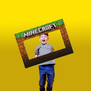 May include: A Minecraft photo frame prop featuring the game's logo and a pixelated earth and grass design. A child in a grey top and blue jeans holds the frame against a yellow backdrop.
