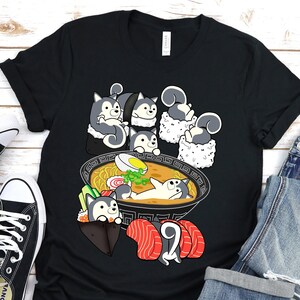 May include: Black t-shirt featuring a cartoon design of dogs in a bowl of ramen. The dogs are wearing sushi hats.